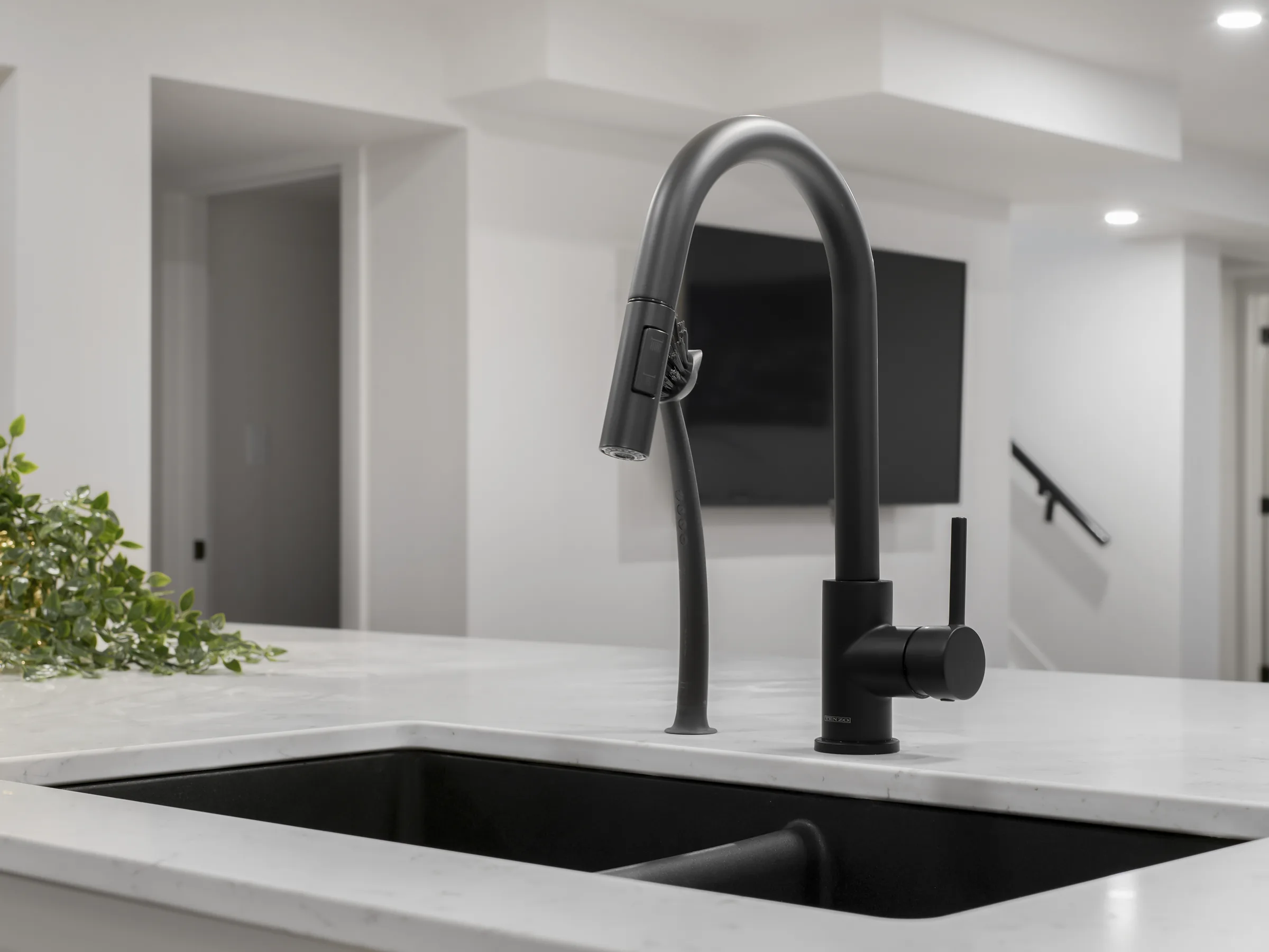 Kitchen faucet close-up over a black sink, TV and stairs visible in the background