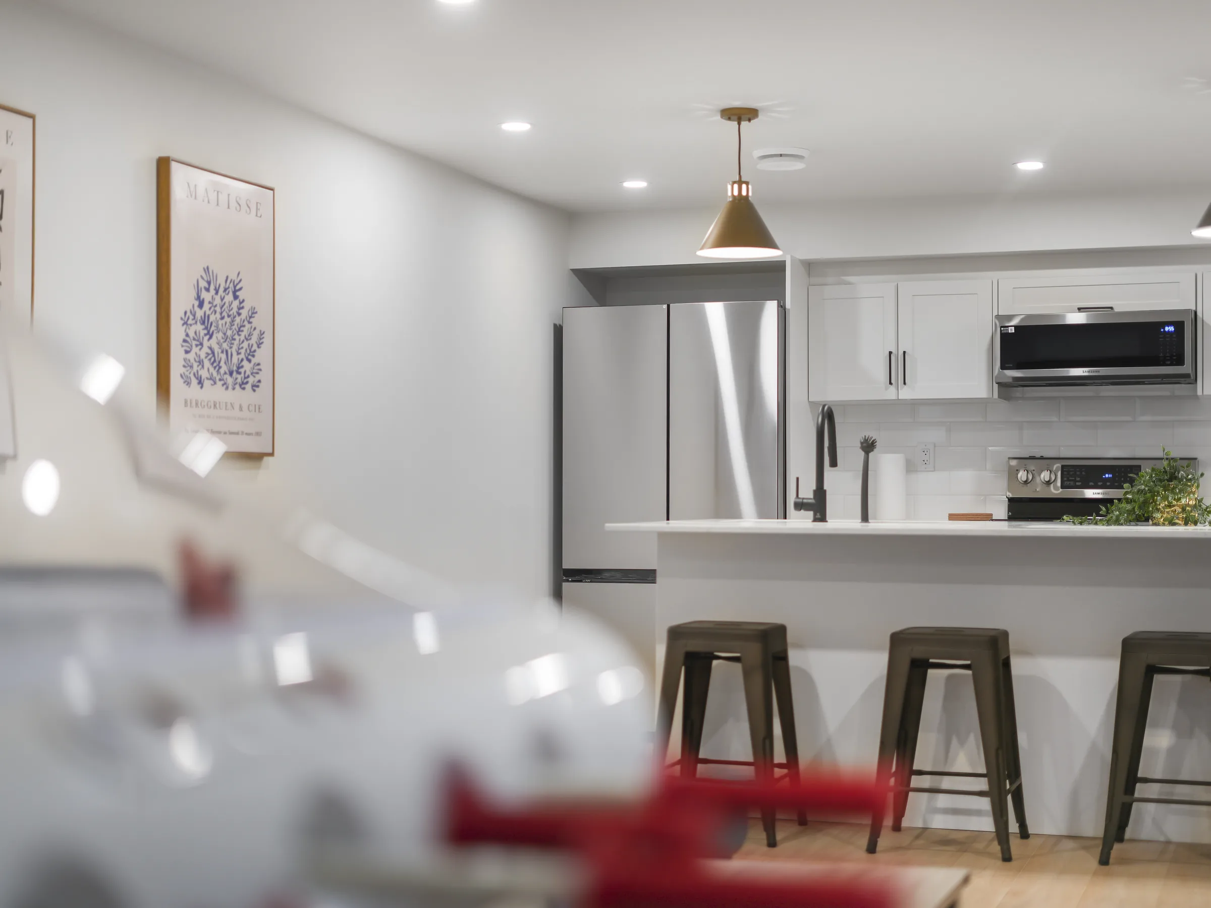 Wide shot through a blurred foreground toward the kitchen, fridge and stools