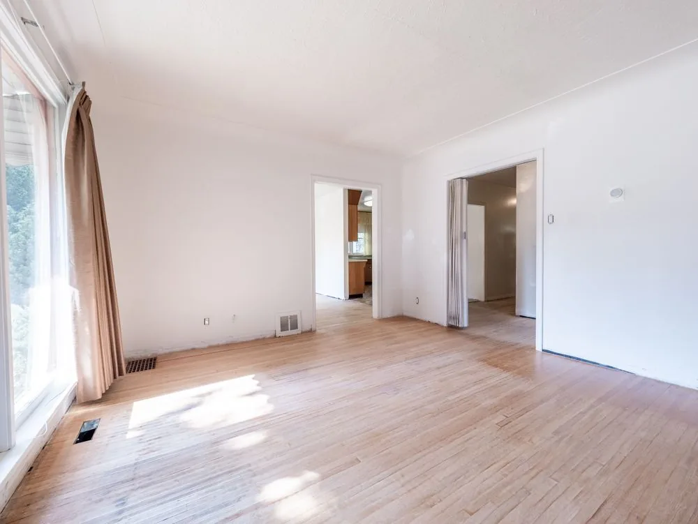 Before renovation, empty living room with window and doorways to the kitchen and a closet