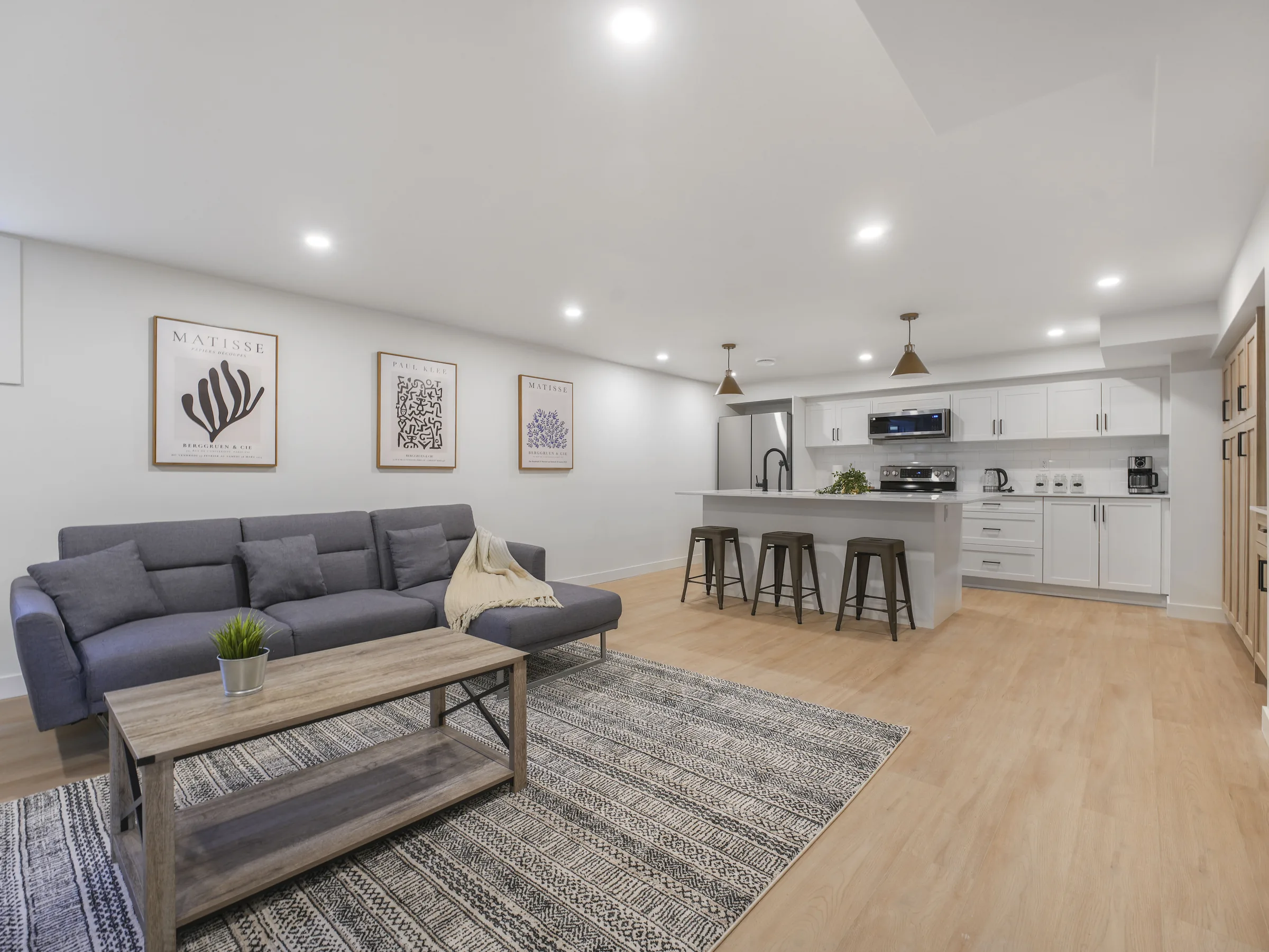 Basement lounge with grey sectional and coffee table, three framed prints on the wall, kitchenette island with three stools beyond