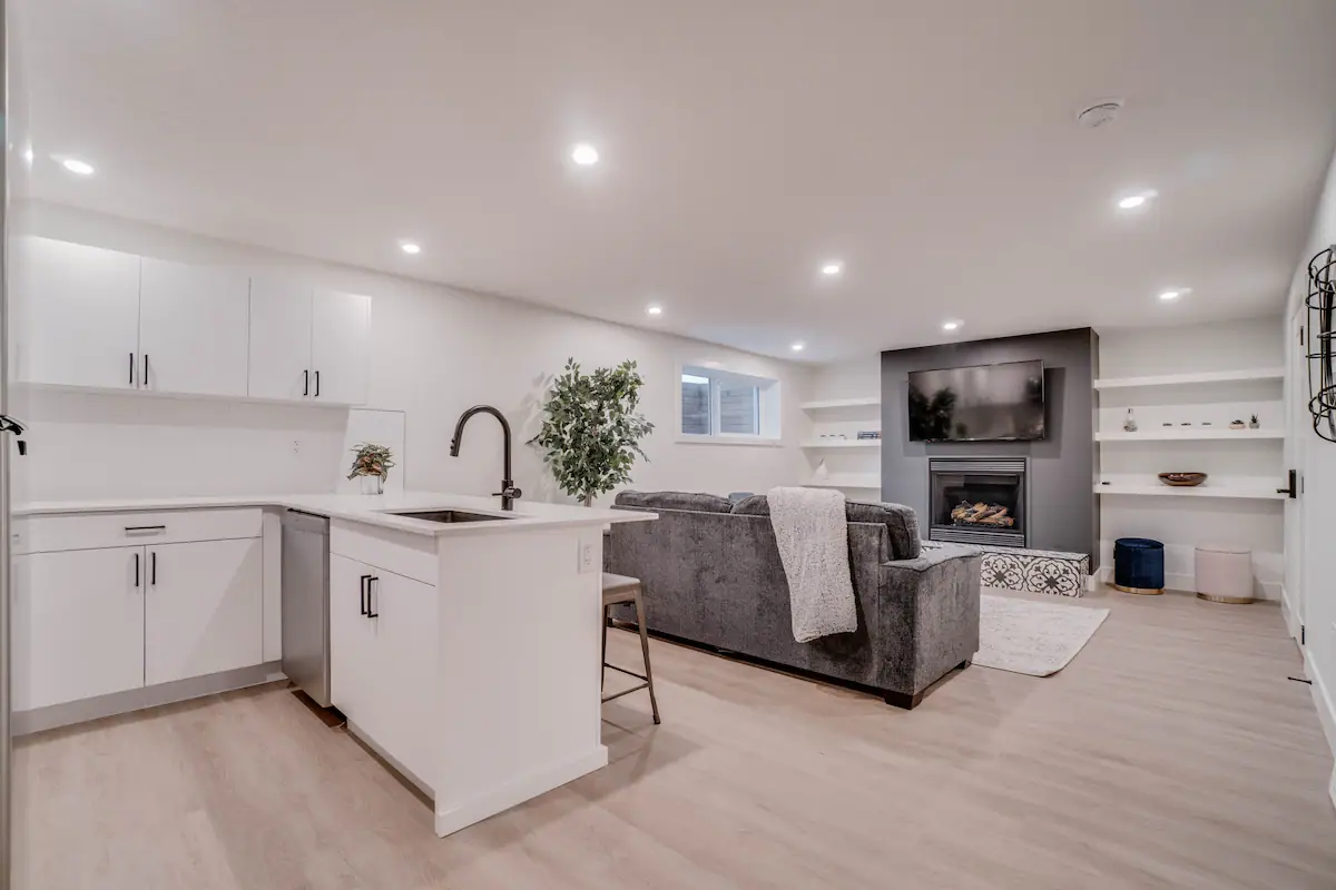Full open plan of the basement suite: white L-shape kitchen with peninsula in the foreground and the fireplace-and-TV living wall beyond.