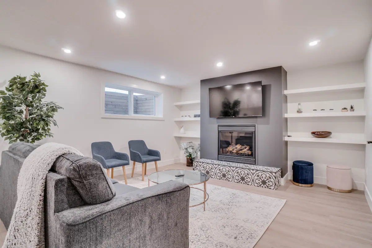Basement suite living room from the opposite corner, showing the built-in floating shelves either side of the fireplace and the egress window at the exterior wall.