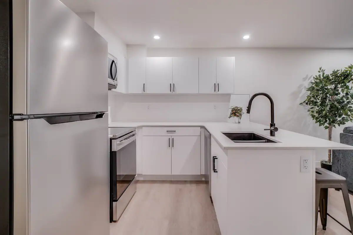 Basement suite kitchen from the far side of the peninsula, showing the stainless fridge, range, and the black gooseneck pull-down faucet at the peninsula sink.