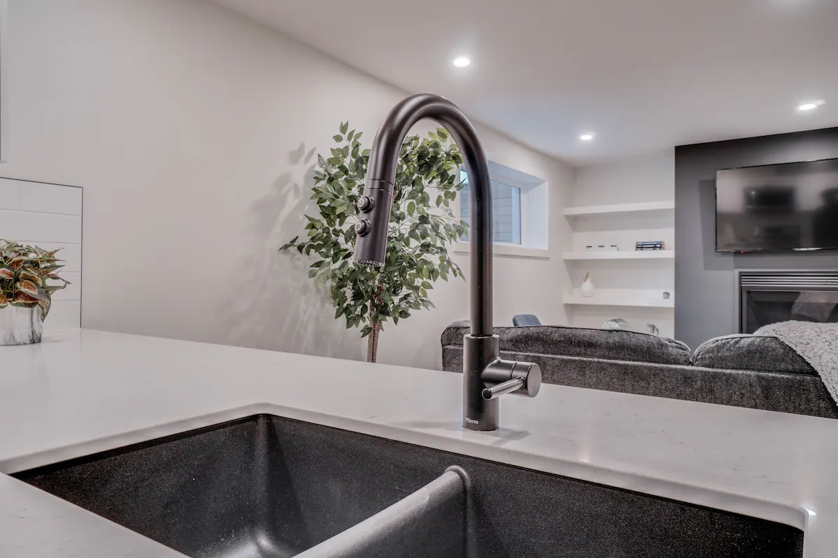 Close-up of the basement suite peninsula sink with black gooseneck pull-down faucet and black composite double basin, with the living room TV and fireplace beyond.