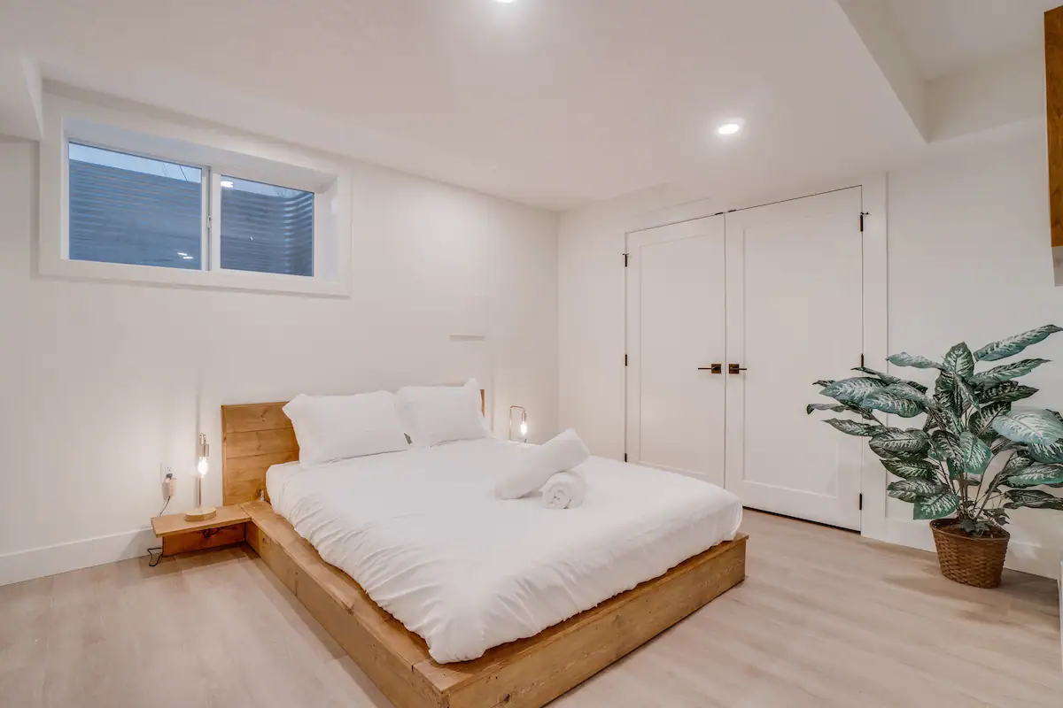 Same basement suite bedroom shown from the foot of the bed, with the double closet doors and a large potted dieffenbachia by the wall.