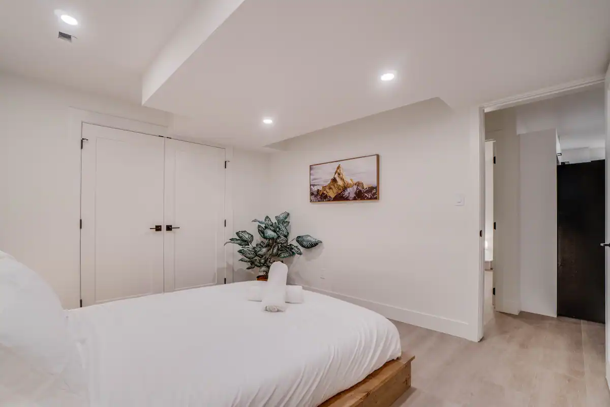 Basement suite bedroom seen from the head of the bed, with the double closet on the left, framed mountain-peak photograph on the far wall, and the hallway opening on the right.