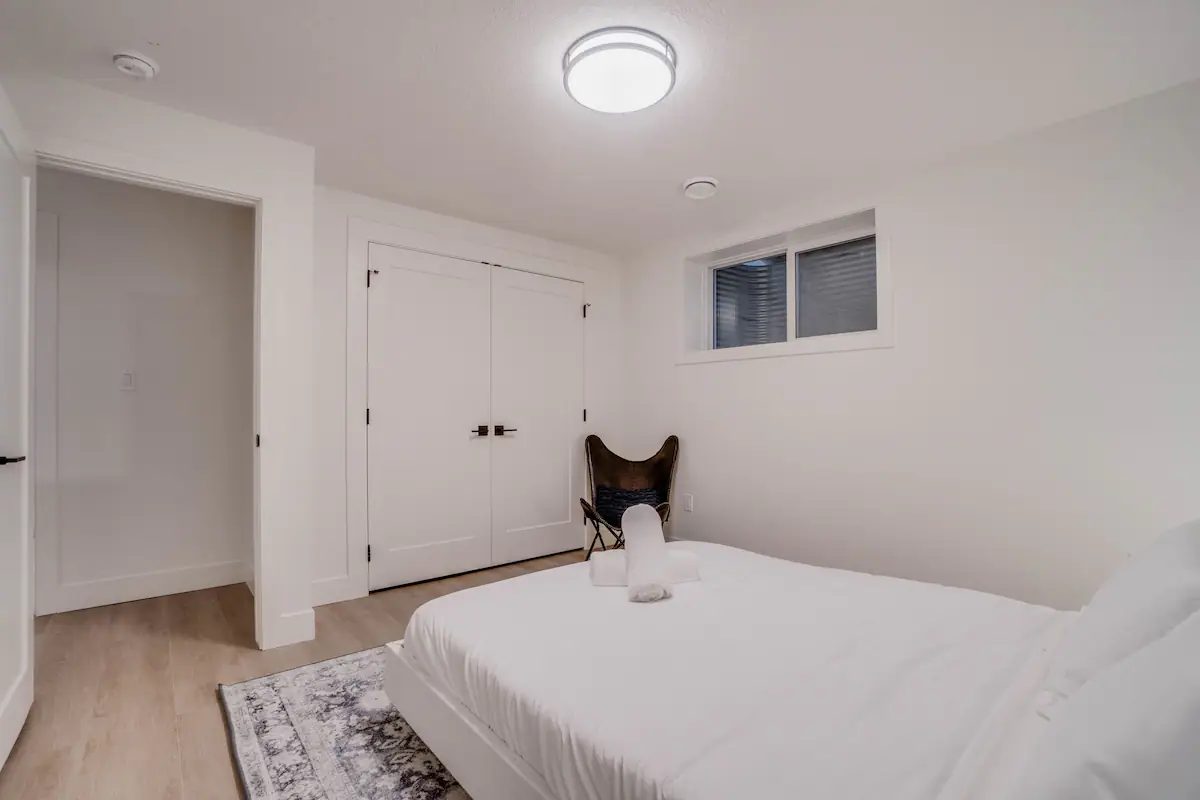 Second basement suite bedroom from the foot, showing the double closet, an egress window above a black leather butterfly chair, and doorway to the hall.