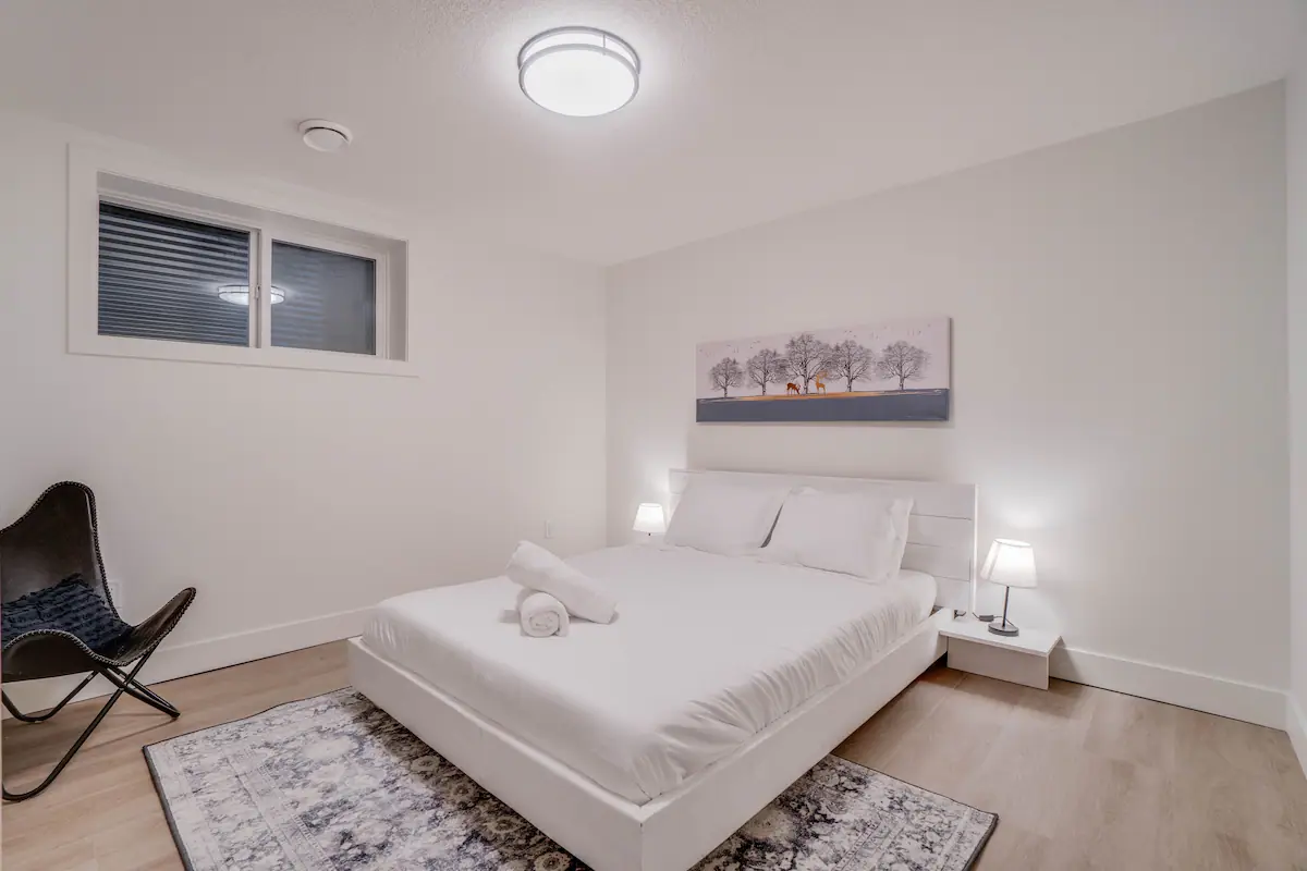 Second basement suite bedroom from the closet corner, showing the bed with matching table-lamp nightstands and the deer-and-trees canvas above.