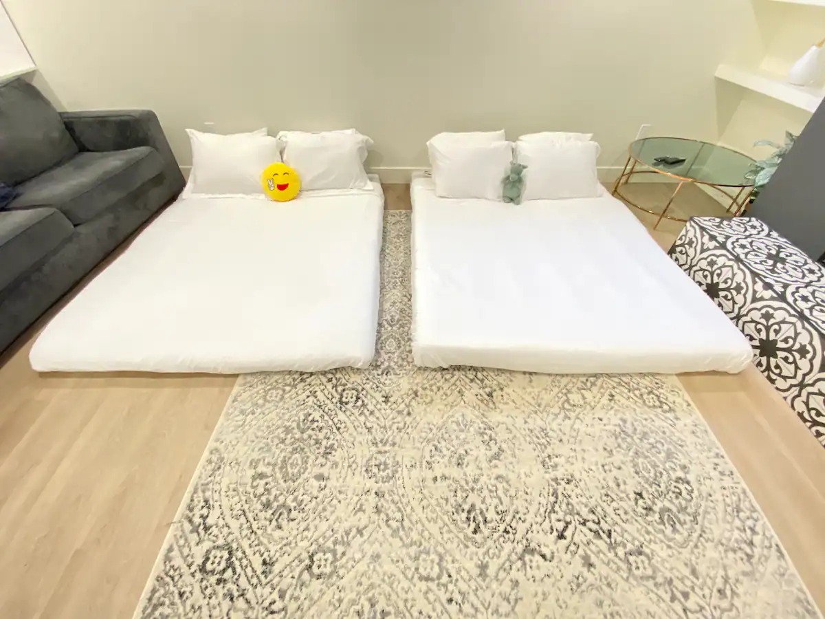 Basement suite living room reconfigured for extra sleeping, with two floor mattresses laid side by side in front of the gray sofa and patterned ottoman bench.