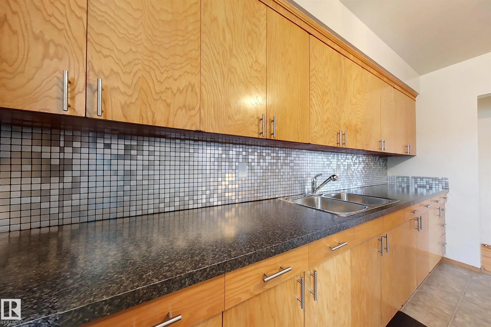 Before renovation, close view of the kitchen countertop, double stainless sink, and silver-glass mosaic-tile backsplash.