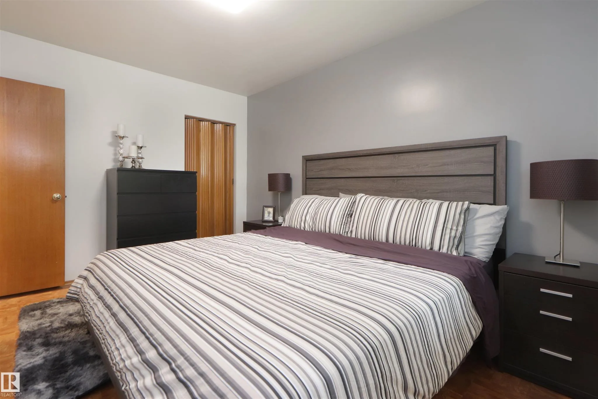 Before renovation, primary bedroom with king bed, gray walls, a wood accordion-style closet, and a small black dresser.