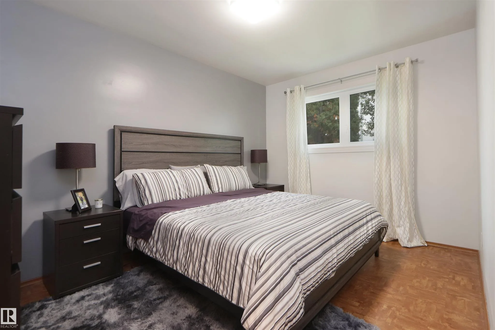 Before renovation, bedroom with a king bed dressed in striped bedding, gray walls, and parquet-pattern flooring.