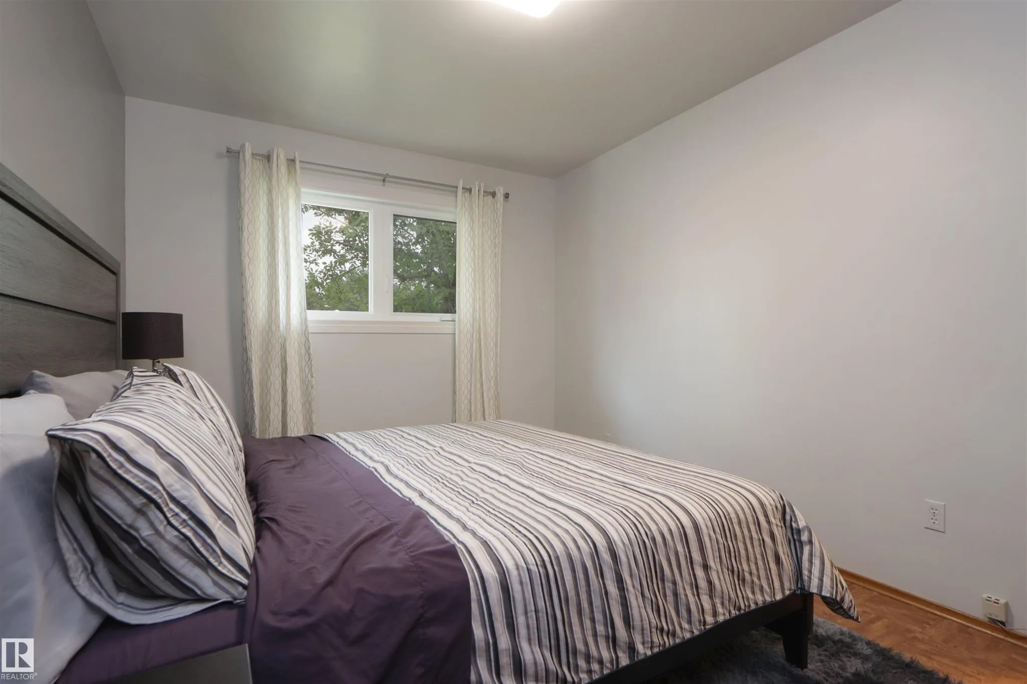 Before renovation, smaller bedroom with a made bed against the wall, drawn curtains, and parquet flooring.