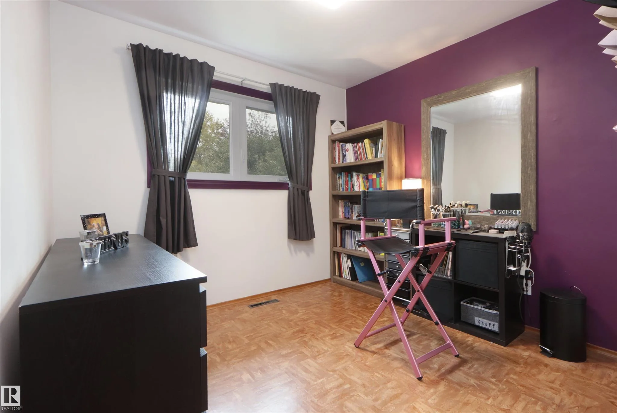 Before renovation, a bedroom in use as a dressing / makeup room with a purple accent wall, wall-mounted mirror, and black wire shelving.