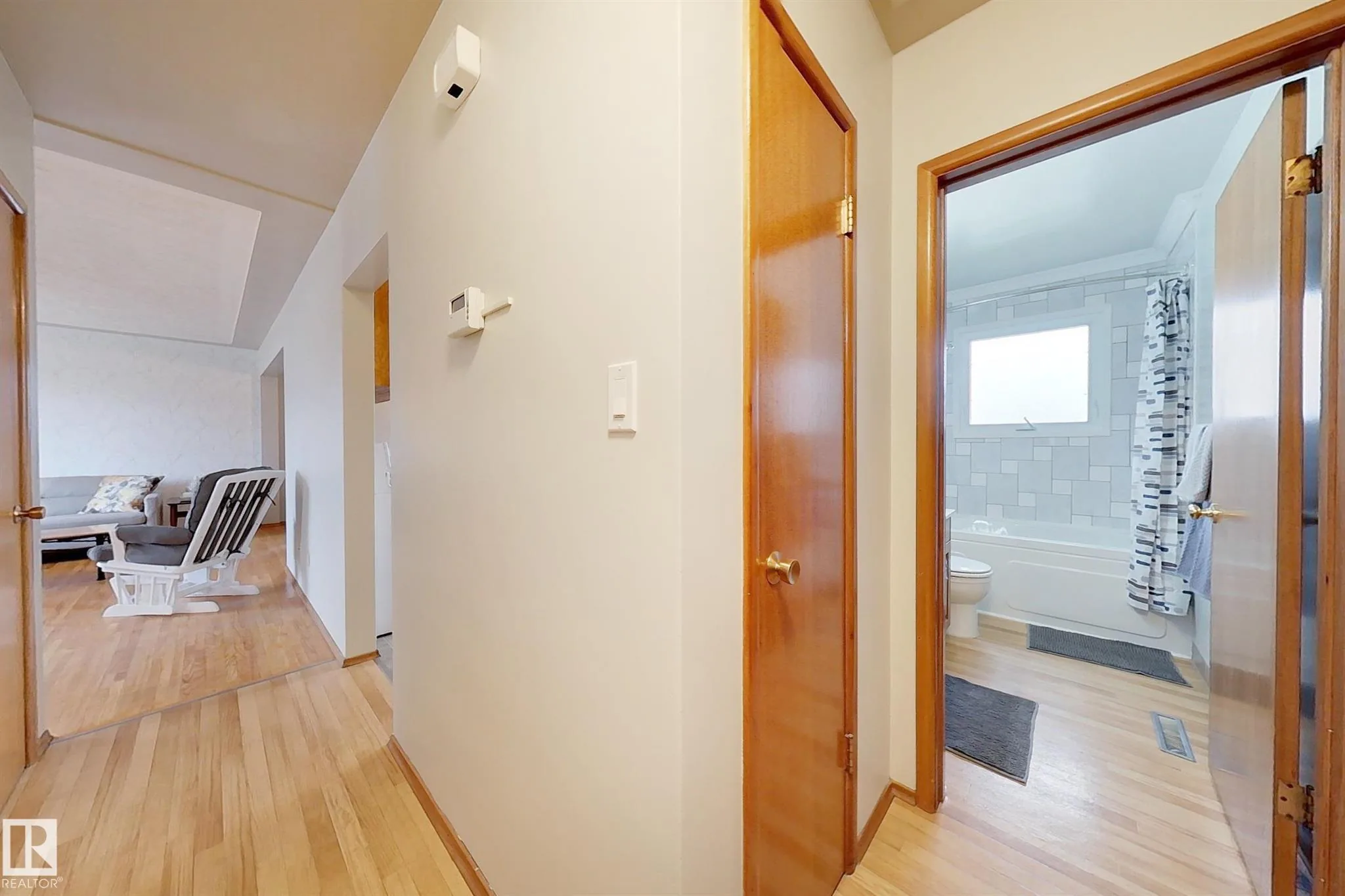 Before renovation, view from the hallway toward the open main-floor bathroom door, showing hardwood floor, thermostat on wall, and glimpse of the subway-tiled tub.