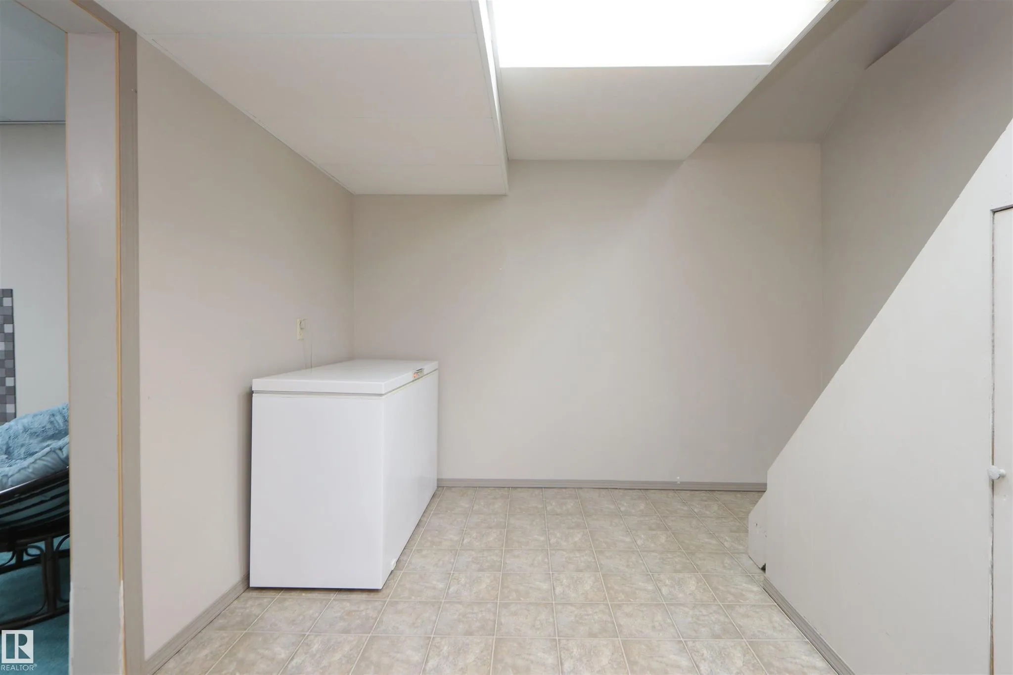 Before renovation, small basement landing / storage area with a white chest freezer against the wall, beige tile floor, and open view under the stairs.