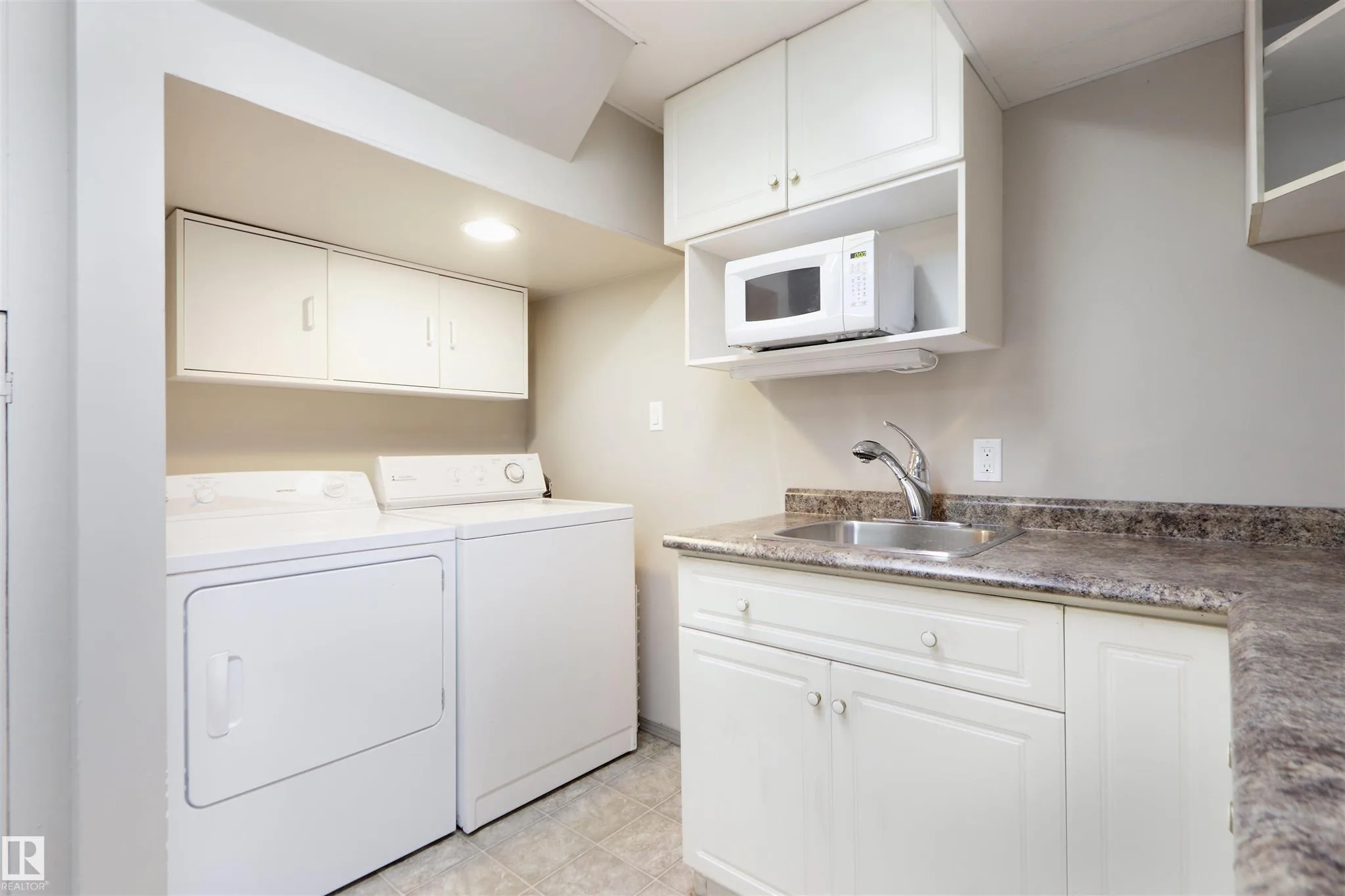 Before renovation, closer view of the basement kitchenette / laundry area showing the stainless sink, wall-mounted white microwave, and side-by-side top-load washer and dryer in the alcove.
