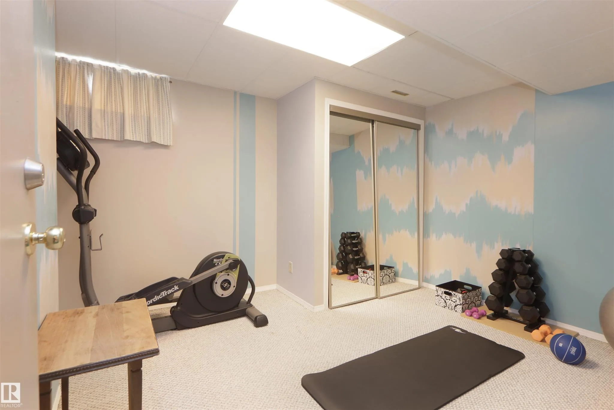 Before renovation, small basement room used as a home gym with NordicTrack elliptical, dumbbells, and a mirrored bifold closet on the tie-dye-painted wall.