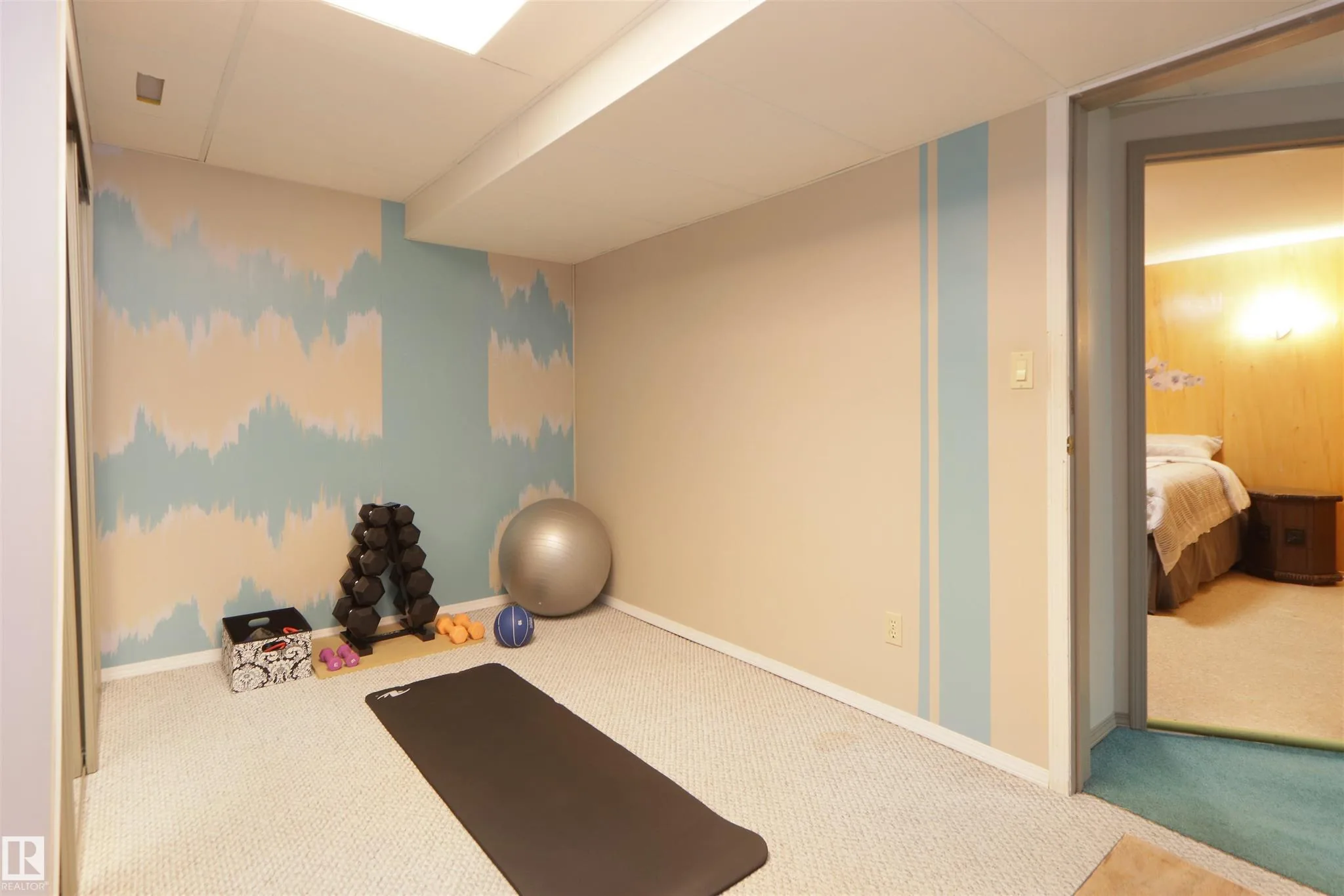 Before renovation, gym room from another angle showing dumbbells and yoga mat on beige carpet, with the door open through to the adjoining basement bedroom.