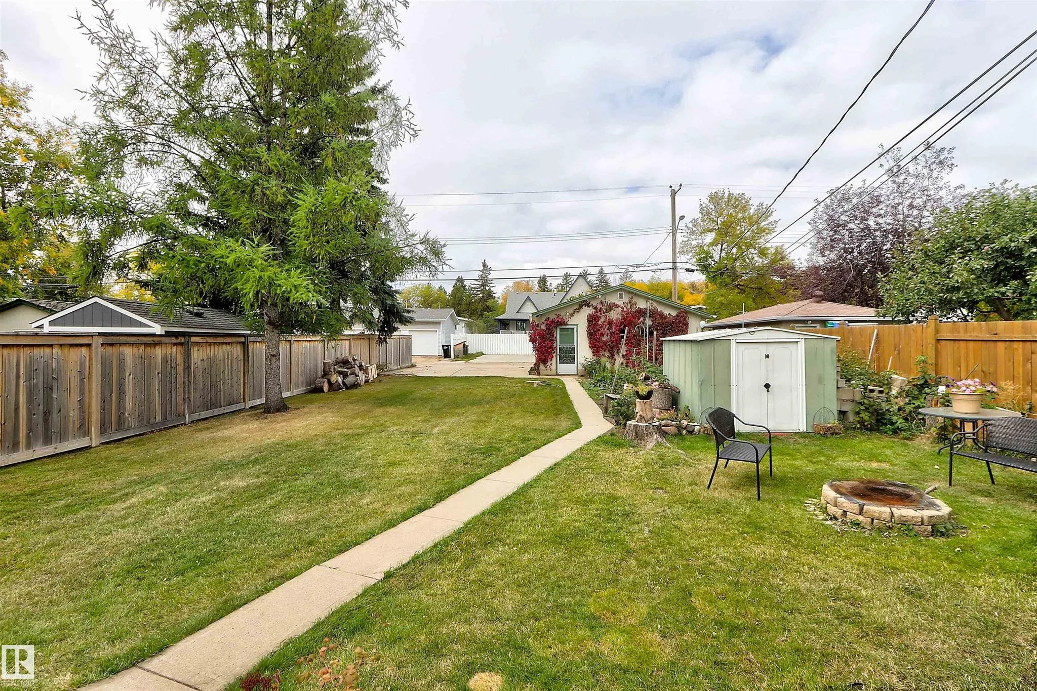 Before renovation, backyard from the concrete path showing green garage door on the detached garage, gray tool shed, and the concrete fire ring seating area.