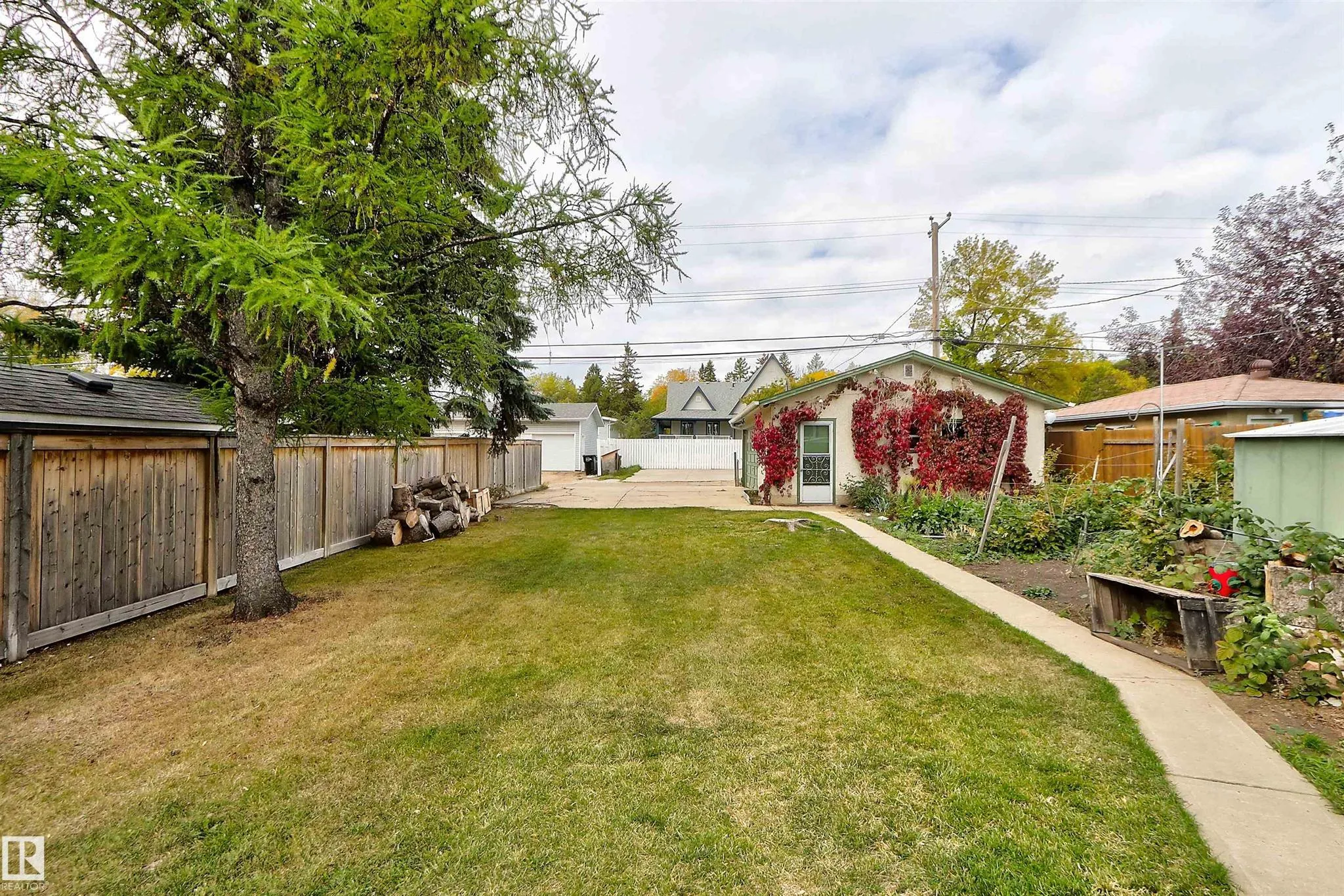 Before renovation, side-yard view down toward the detached garage's back door with vegetable garden along the right fence and firewood stacked against the left fence.