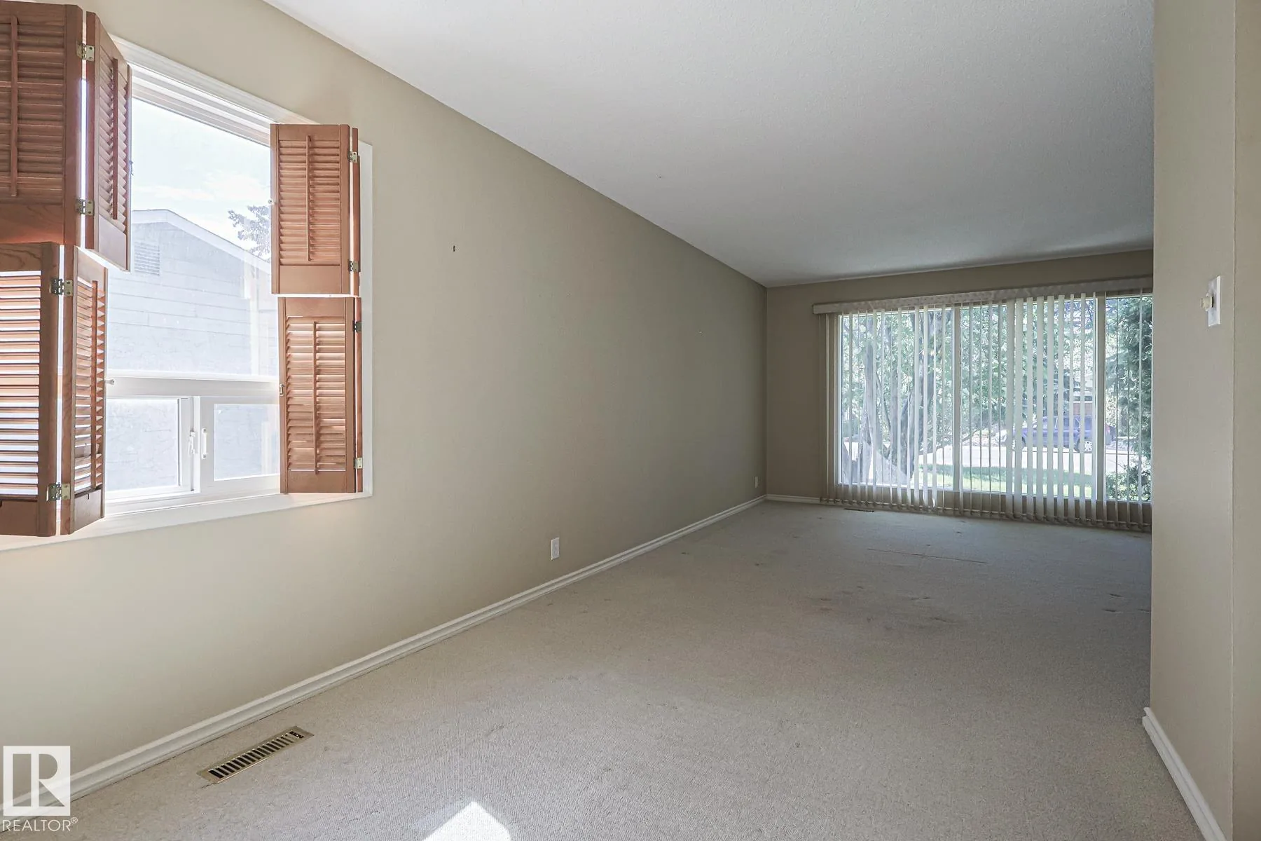 Before renovation, a corner of the living room showing folding wooden louver shutters open on a smaller side window and vertical blinds on the larger picture window.