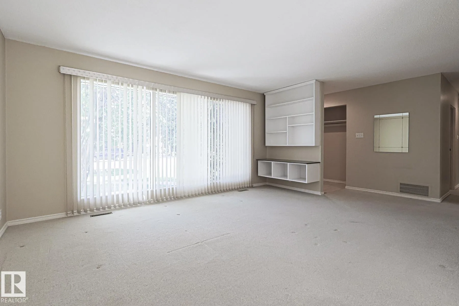 Before renovation, the empty living room with vertical blinds on the large front window and a built-in white shelf with a low storage bench.