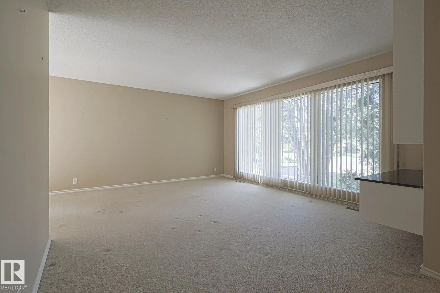 Before renovation, a wide empty living room with beige carpet and vertical blinds on the large front window; kitchen counter with dark laminate top visible at right.