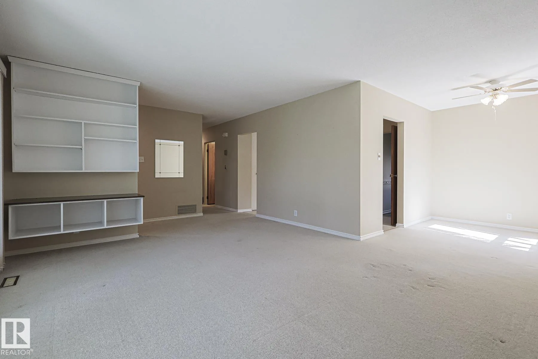 Before renovation, view across the living room toward a built-in white shelf-and-storage unit and hallway leading to bedrooms and bath.