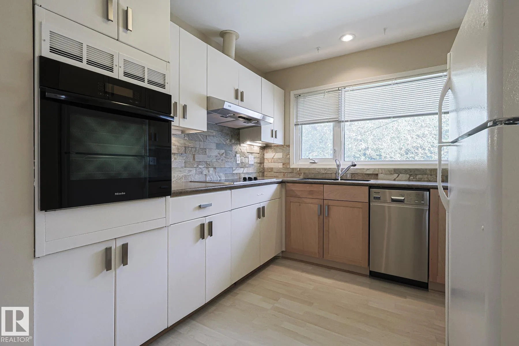 Before renovation, the kitchen with two-tone cabinetry (white uppers, wood shaker lowers), a built-in Miele wall oven, stainless dishwasher, and multi-tone stacked-stone backsplash.