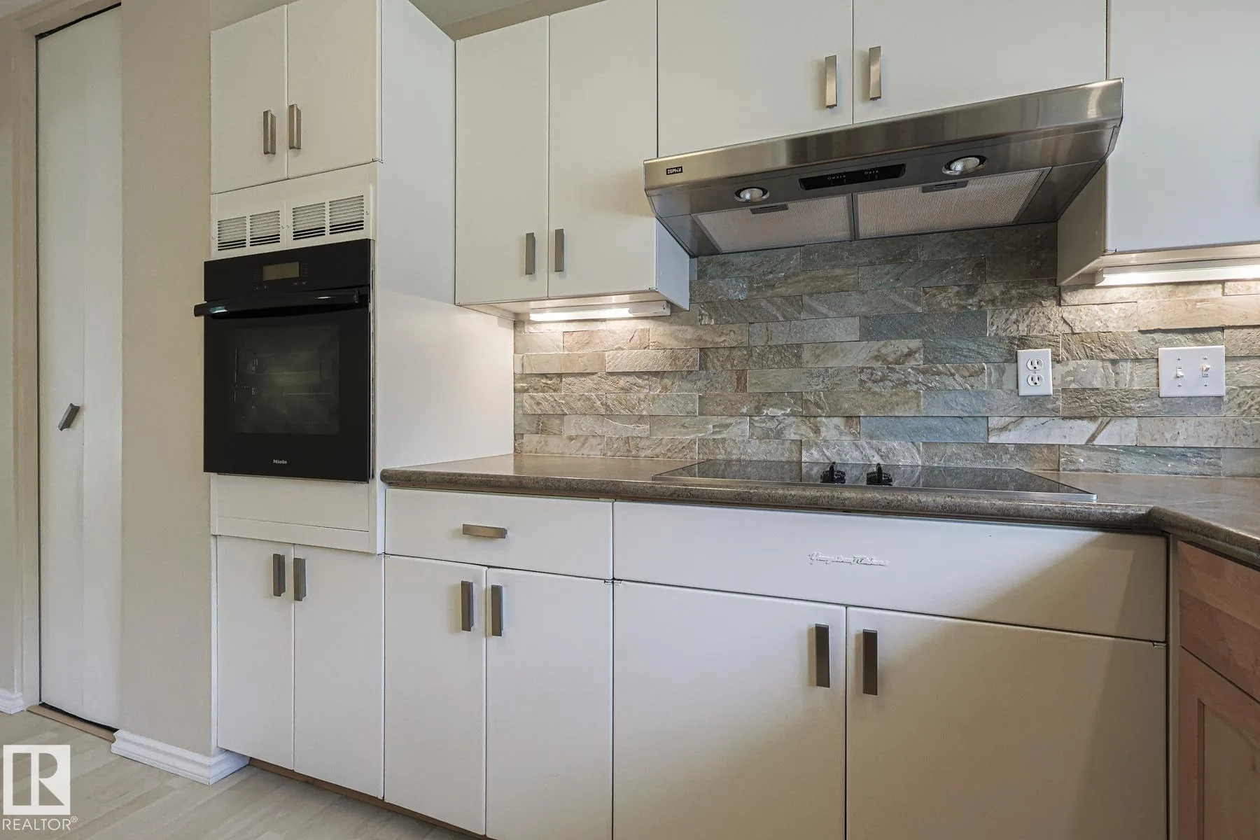 Before renovation, kitchen showing the built-in Miele oven wall with white uppers, stainless range hood, glass cooktop and stacked-stone backsplash.