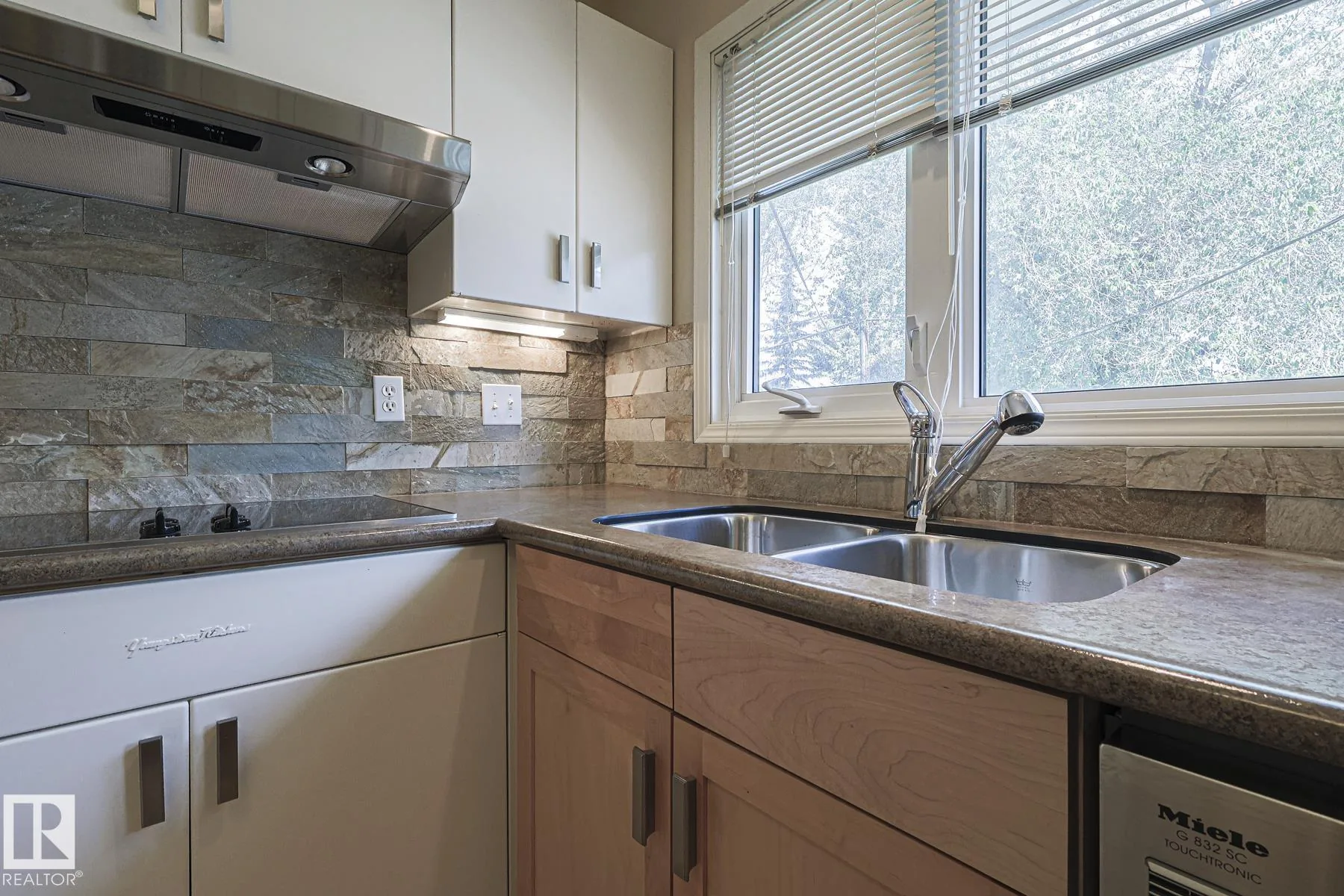 Before renovation, the kitchen sink corner with stainless double-basin sink, chrome pull-out faucet, blinds on the window over the sink, and stacked-stone backsplash.