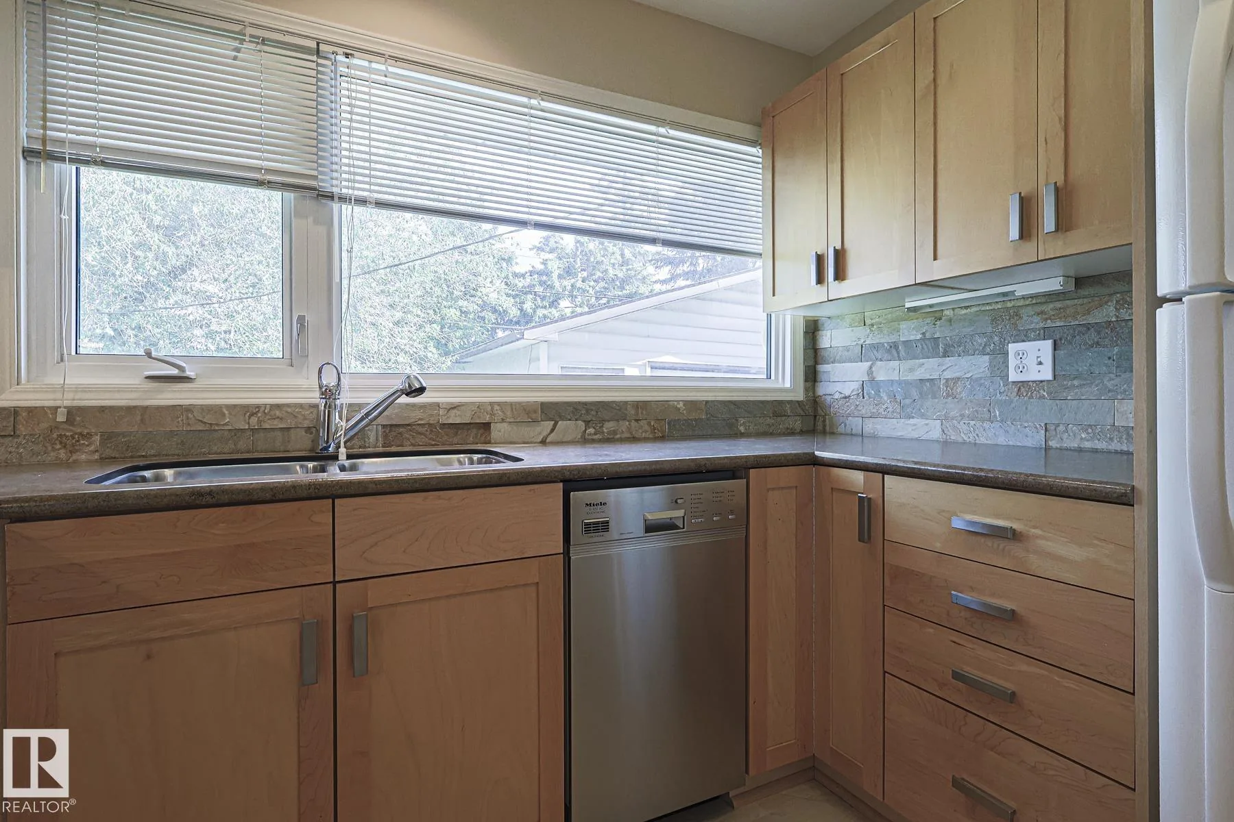 Before renovation, kitchen sink wall from a different angle showing wood shaker lower cabinets, the stainless dishwasher, and the same stacked-stone backsplash.
