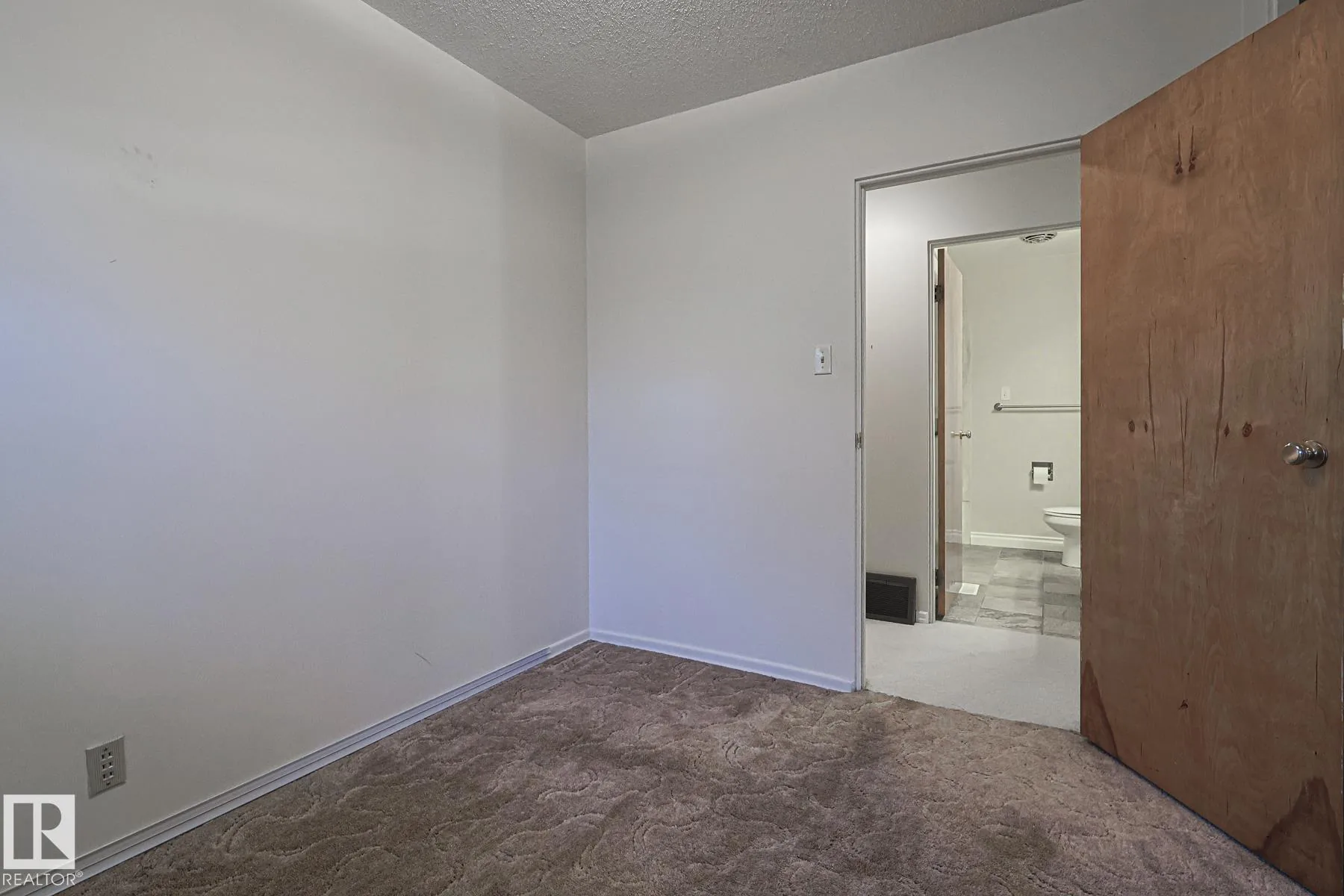 Before renovation, a bedroom with brown patterned carpet and a plywood door, with a main-floor bathroom visible through the open doorway.