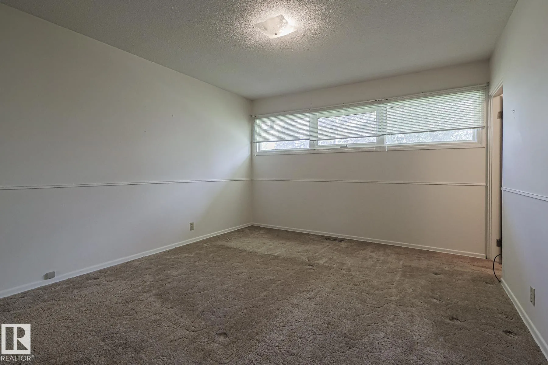 Before renovation, a small bedroom with brown patterned carpet, a chair-rail trim line, and a wide horizontal window near the ceiling.