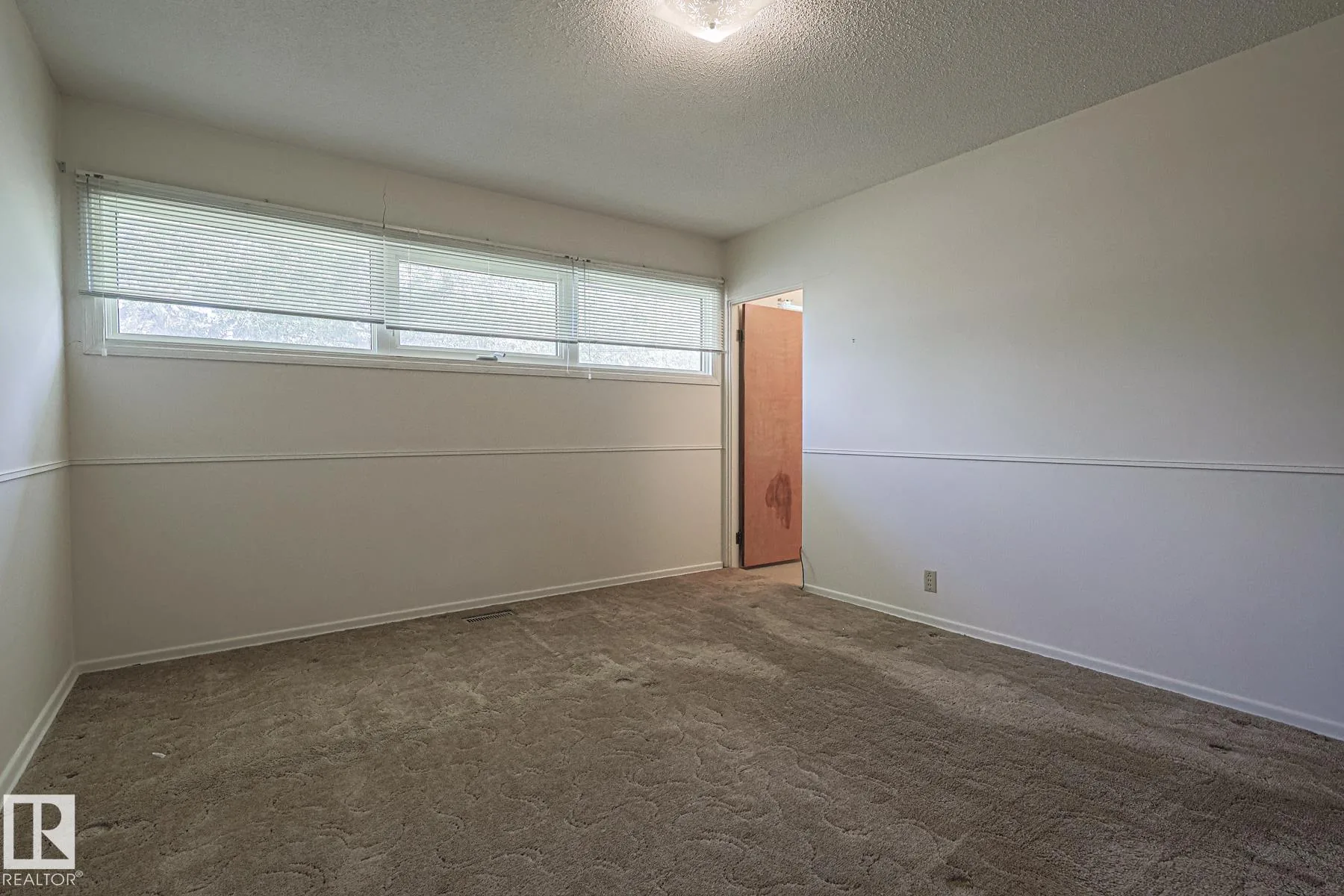Before renovation, a bedroom with brown patterned carpet, chair-rail trim, a plywood swing door, and a narrow horizontal window near the ceiling.