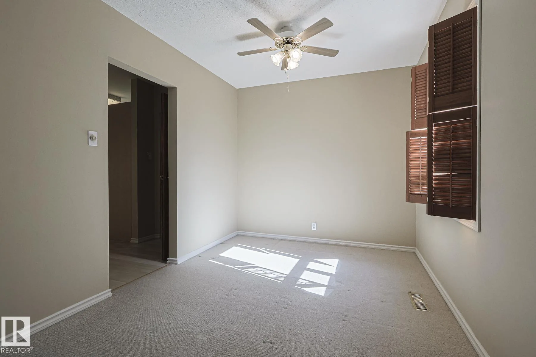 Before renovation, a bedroom with beige carpet, brown wood folding louver shutters on the window, and a wood ceiling fan with light kit.