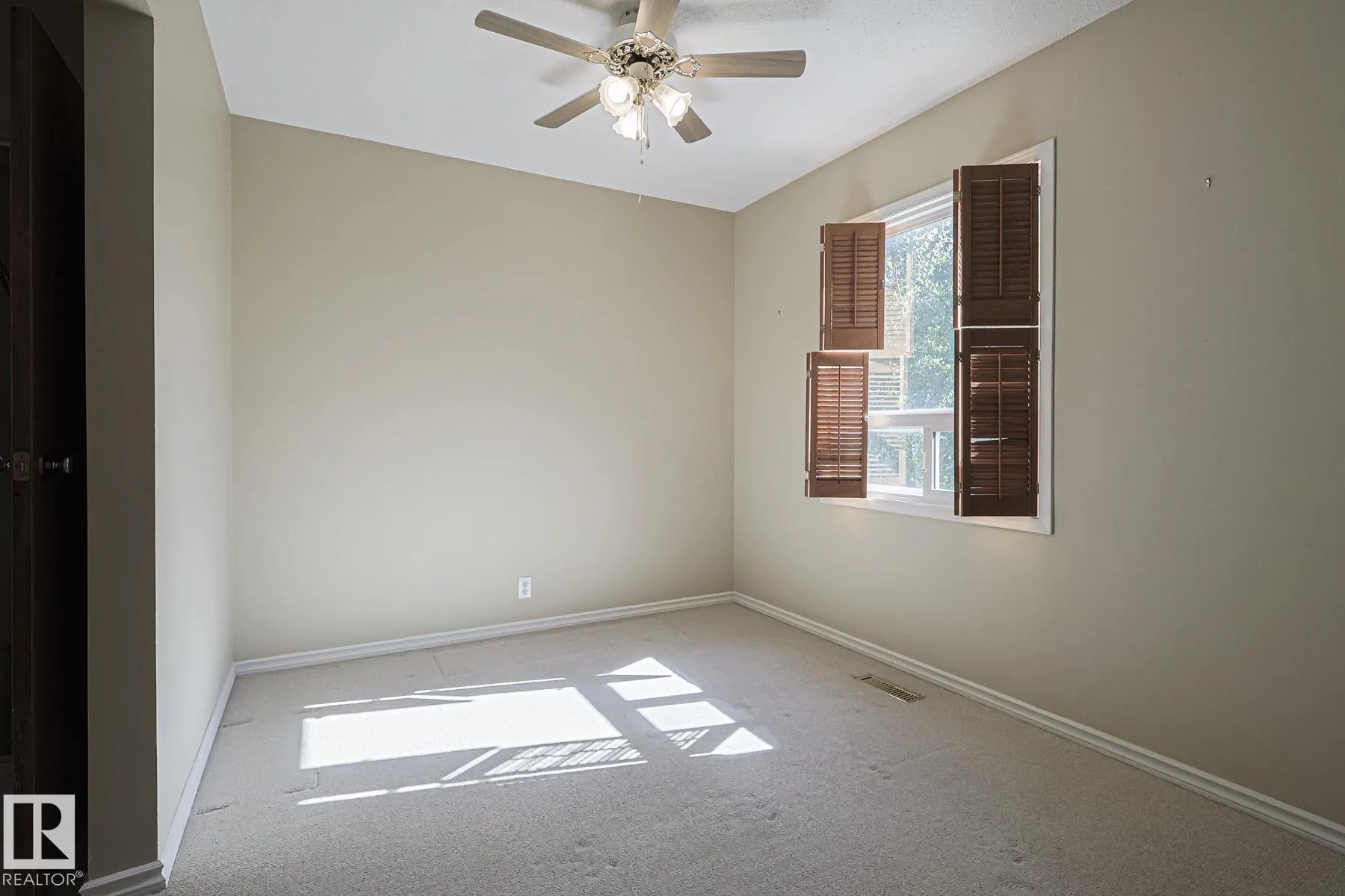 Before renovation, a larger bedroom with beige carpet, ceiling fan, and a folding wooden louver shutter on the window at back wall.