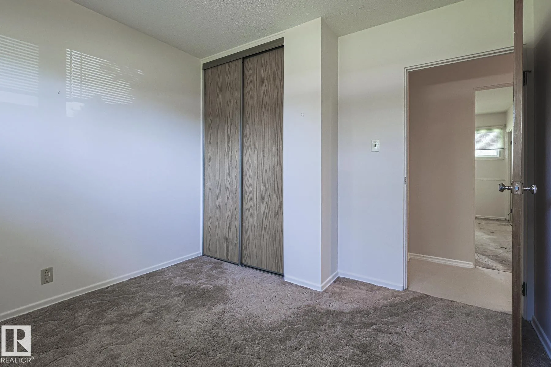 Before renovation, a bedroom with brown patterned carpet, a woodgrain sliding closet, and a plywood door open to the hallway.