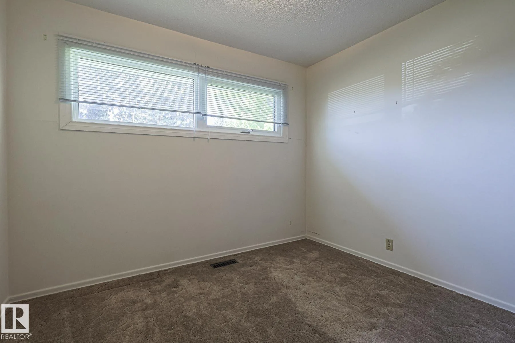 Before renovation, a small bedroom with brown patterned carpet and wide horizontal windows with white blinds on two walls.