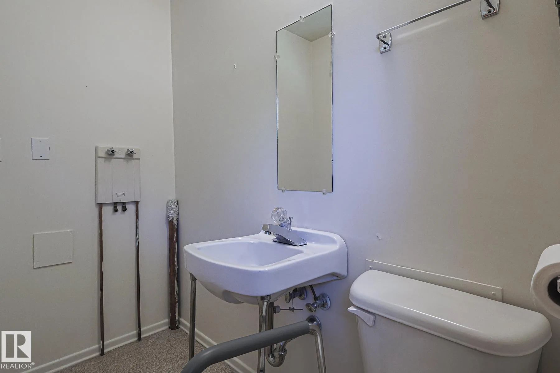 Before renovation, a bathroom with a wall-mounted sink on chrome legs, two rectangular wall mirrors, and a toilet with a grab-bar frame.