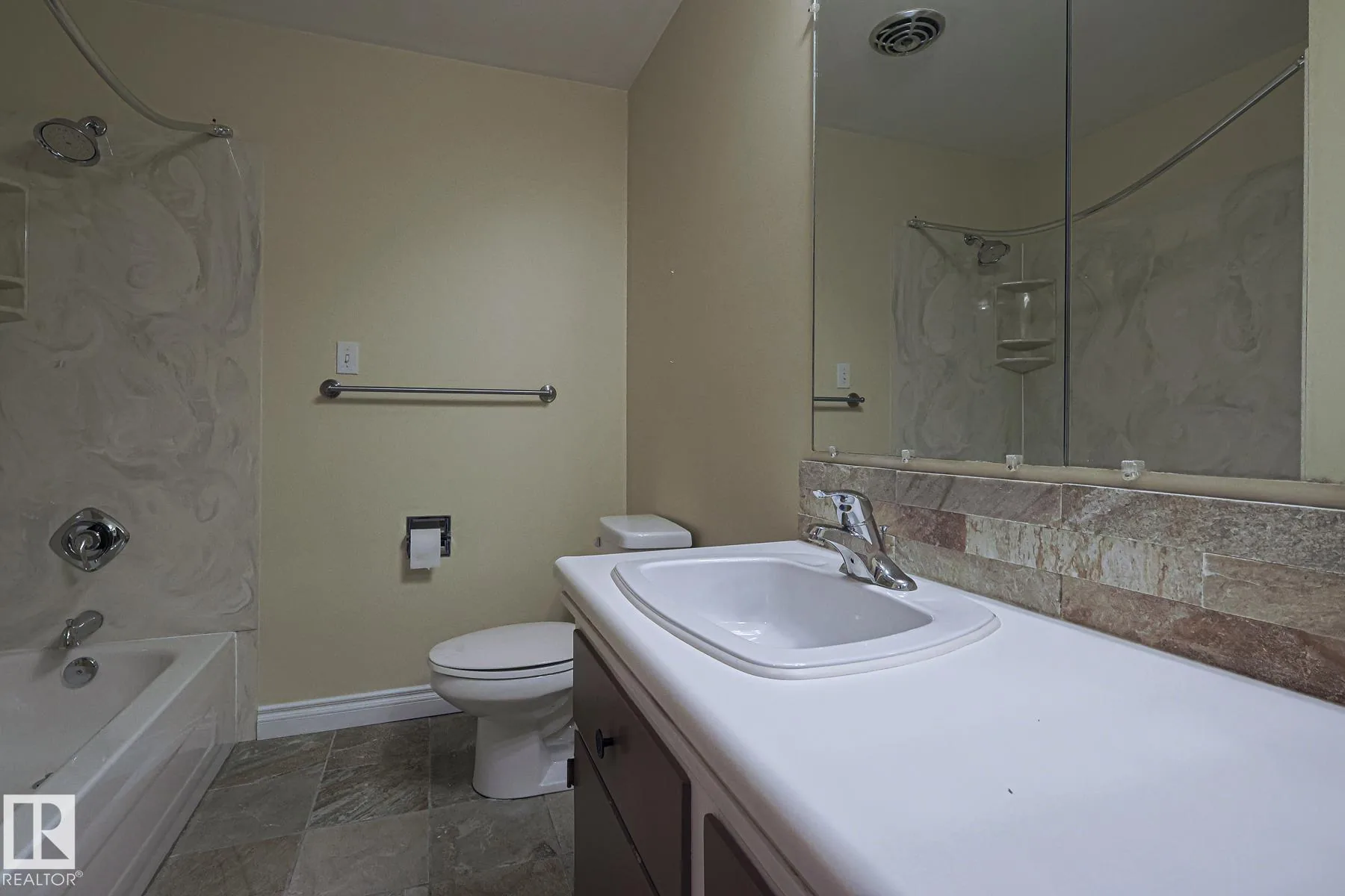 Before renovation, a full bathroom with an alcove tub-shower under a curved shower rod, marble-swirl surround, dark vanity with white top, and slate-look tile floor.