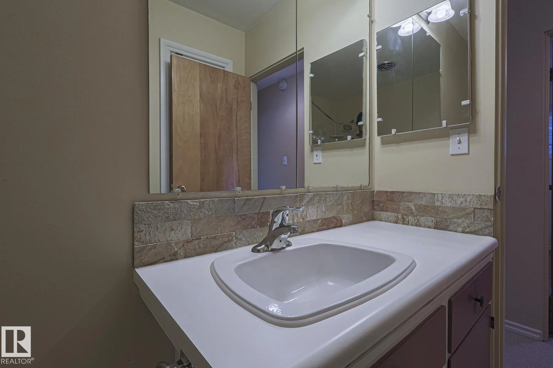 Before renovation, a bathroom vanity with a white drop-in sink, dark laminate counter, stacked-stone backsplash strip, and mirrored medicine cabinets above.