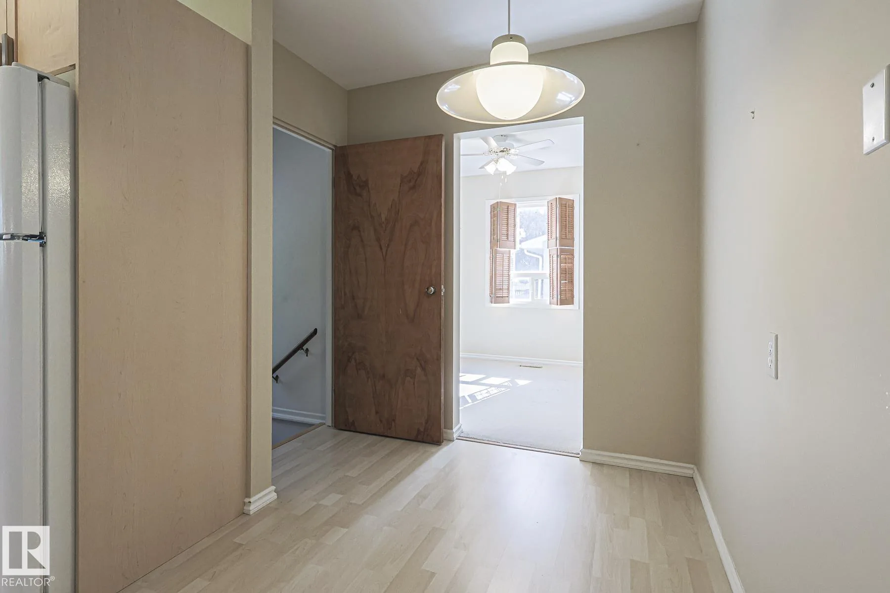 Before renovation, a small eat-in nook next to the kitchen with pale-wood laminate floor, a modern white bell pendant lamp, and a plywood door open toward the adjacent living area.