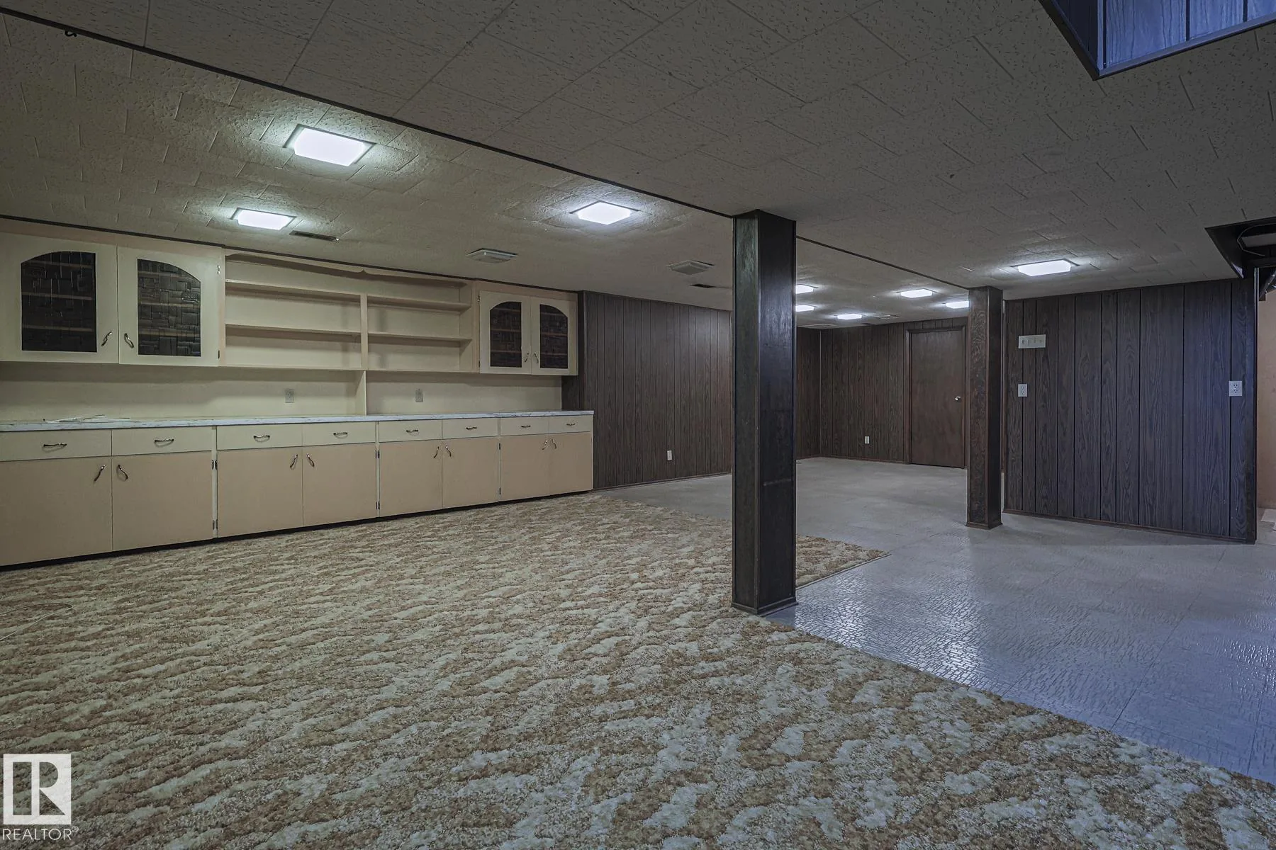 Before renovation, the basement rec area with cream built-in cabinets on the left wall, dark faux-wood paneling on the columns, and the shag carpet zone leading to a further paneled area.