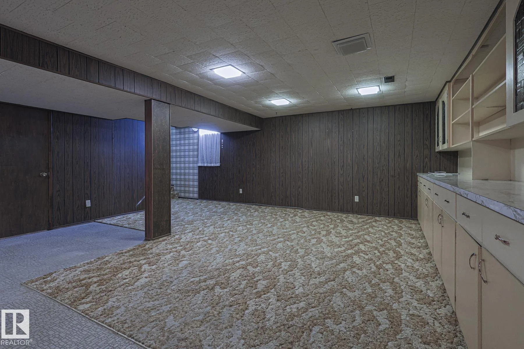 Before renovation, the basement rec area showing another angle of the cream built-in cabinetry, dark paneled walls, and a small hopper window with sheer curtain.