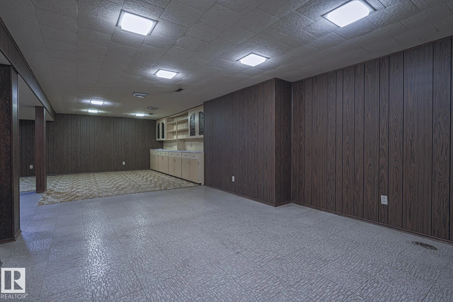 Before renovation, wide view of the basement showing the vinyl-tile floor, dark faux-wood paneling, drop-tile ceiling, and the cream built-in cabinet run in the middle distance.