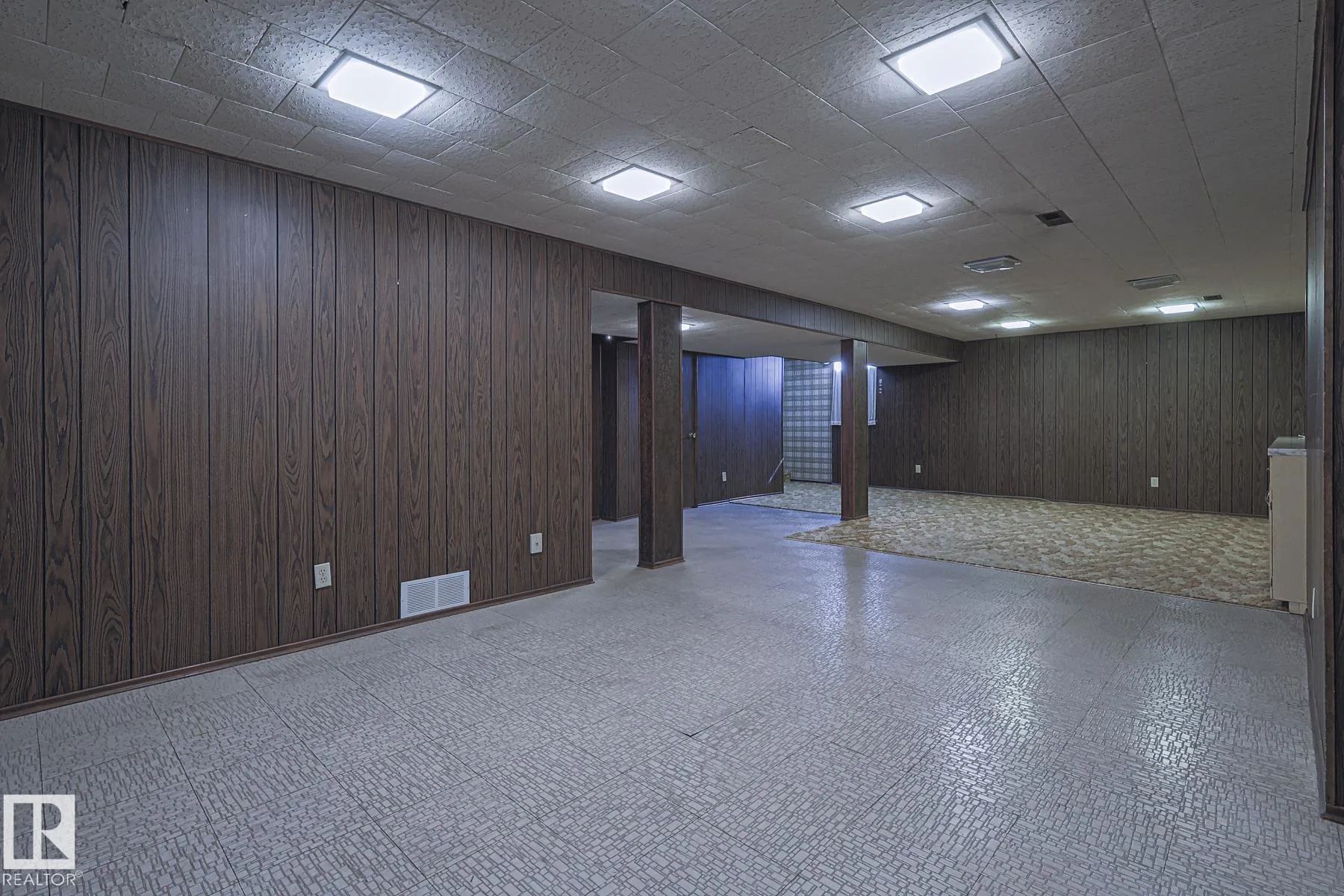 Before renovation, basement space with dark faux-wood paneling, drop-tile ceiling, and a shag carpet zone visible through the boxed column opening at right.
