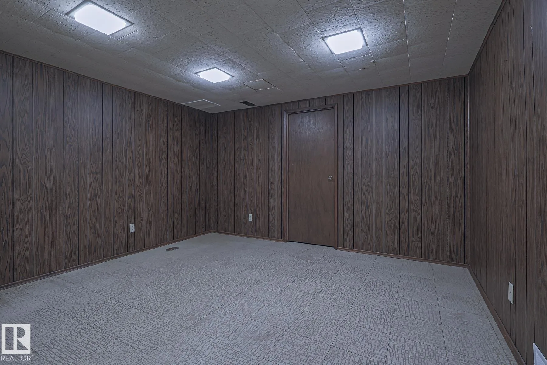 Before renovation, a small windowless basement room with dark faux-wood paneled walls, drop-tile ceiling, a dark-stained door, and vinyl parquet-pattern floor tile.