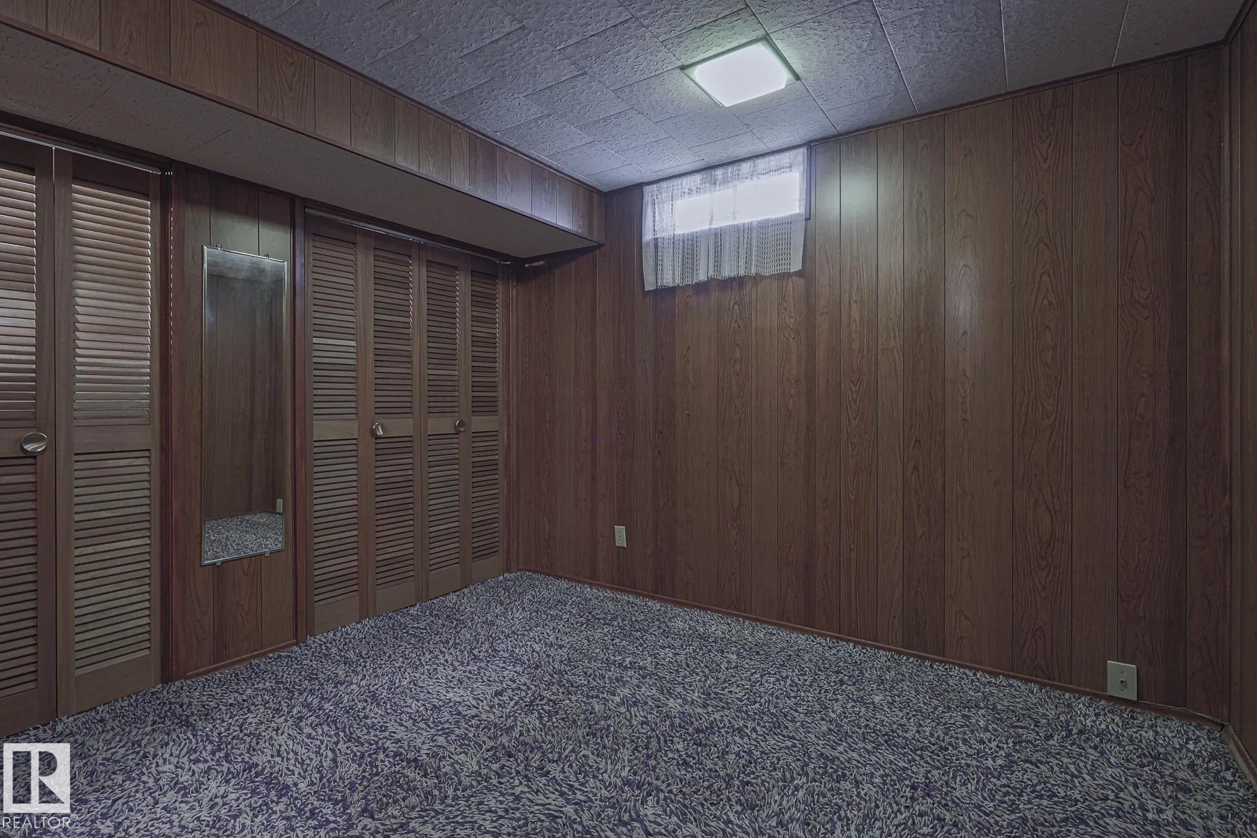 Before renovation, a basement bedroom with dark faux-wood paneling, purple-and-cream shag carpet, a small hopper window with sheer curtain, and a run of louvered closet doors along one wall.